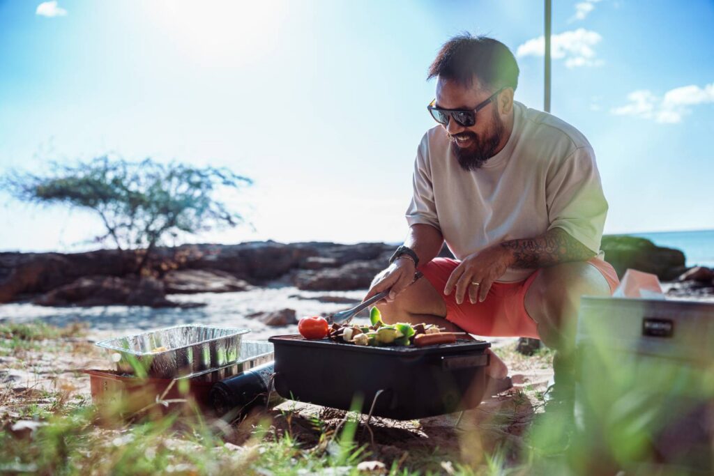 A man of Asian descent squats in the sand at the beach and grills hot dogs and vegetable kabobs on a portable barbecue while enjoying a relaxing day at the beach with family and friends.